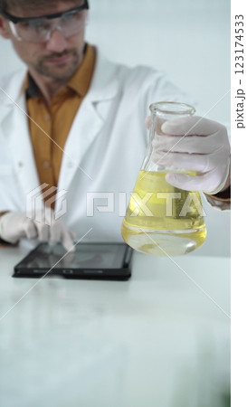 Man scientist wearing white protective gloves, and glasses is holding a yellow chemical solution inside an Erlenmeyer flask in a laboratory, vertical portrait Man scientist wearing white protective gloves, and glasses is holding a yellow chemical solution inside an Erlenmeyer flask in a laboratory, vertical portrait 123174533