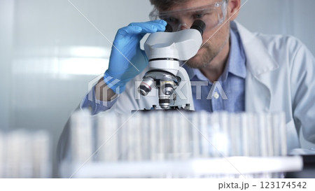 Male scientist researcher wearing blue gloves, protective glasses is using microscope in modern laboratory, portrait view. Low key lighting photo. Science and Medicine concepts Male scientist researcher wearing blue gloves, protective glasses is using microscope in modern laboratory, portrait view. Low key lighting photo. Science and Medicine concepts 123174542