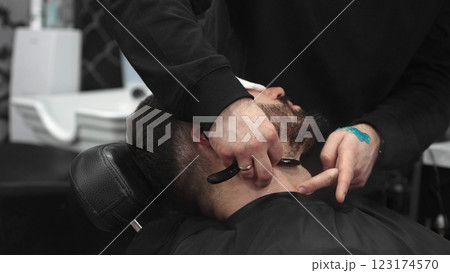 Professional barber carefully shaving client's beard with straight razor, applying gel and using hot towel for precise grooming in contemporary barbershop setting Professional barber carefully shaving client's beard with straight razor, applying gel and using hot towel for precise grooming in contemporary barbershop setting 123174570