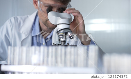 Male scientist wearing lab coat and protective glasses using microscope doing research in modern laboratory setup, portrait view. Low key lighting photo. Science and Medicine concept 123174571