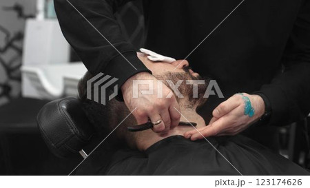 Professional barber carefully shaving client's beard with straight razor, applying gel and using hot towel for precise grooming in contemporary barbershop setting 123174626