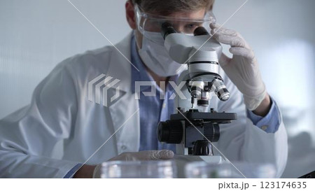 Man scientist researcher wearing white gloves, protective glasses and medical mask is using microscope in modern laboratory, portrait view. Low key lighting photo. Science and Medicine 123174635