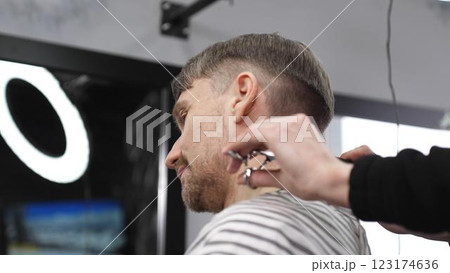 Professional barber trimming blond heard male client's hair with scissors and comb, creating precise modern hairstyle inside contemporary grooming salon 123174636