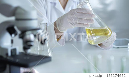 Microscope is near a scientist, wearing a lab coat and white protective gloves, and holding beaker with a yellow oily liquid in laboratory setting, close up. Medicine and science concept Microscope is near a scientist, wearing a lab coat and white protective gloves, and holding beaker with a yellow oily liquid in laboratory setting, close up. Medicine and science concept 123174652