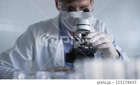 Man scientist researcher wearing white gloves, protective glasses and medical mask is using microscope in modern laboratory, portrait view. Low key lighting photo. Science and Medicine 123174653