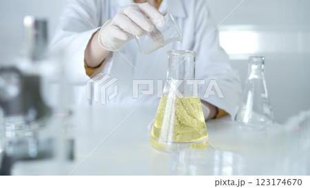 A scientist, wearing a lab coat and white protective gloves, is pouring a yellow oily liquid from one beaker to another in laboratory, close up. Medicine and science concept A scientist, wearing a lab coat and white protective gloves, is pouring a yellow oily liquid from one beaker to another in laboratory, close up. Medicine and science concept 123174670