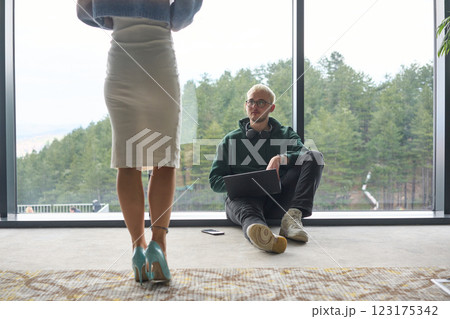 A blonde man sitting casually on the floor of a modern office, leaning against a window as he uses his laptop, with his attractive female colleague standing nearby 123175342