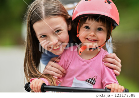 A close up of a teenage girl warmly hugging a young girl wearing a protective helmet while riding a bike in a natural outdoor setting 123175399
