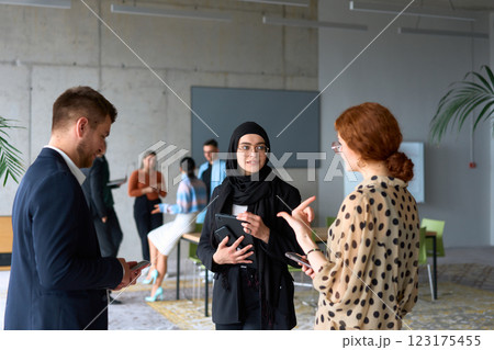 A diverse group of business professionals engages in a conversation during a work break in the office, with their colleagues visible in the background, fostering collaboration and connection 123175455