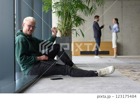 A casual young professional sitting by a large window in a modern office, smiling while working on a laptop, with colleagues interacting in the blurred background. 123175484