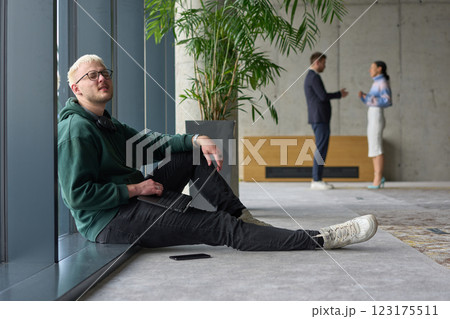 A casual young professional sitting by a large window in a modern office, smiling while working on a laptop, with colleagues interacting in the blurred background. A casual young professional sitting by a large window in a modern office, smiling while working on a laptop, with colleagues interacting in the blurred background. 123175511