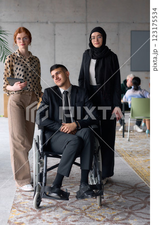A diverse business team poses confidently, including a smiling director seated in a wheelchair, flanked by two professional female colleagues, one wearing a hijab and the other holding a tablet, in a 123175534
