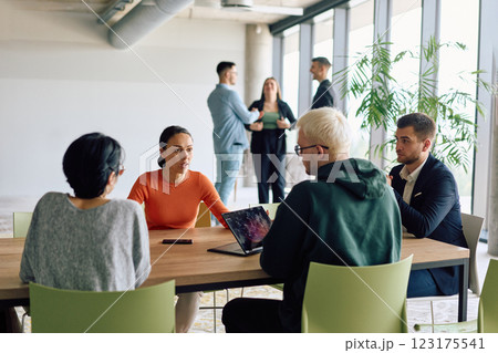 A diverse team of business professionals engaged in a discussion around a conference table in an office, while their colleagues collaborate in the background A diverse team of business professionals engaged in a discussion around a conference table in an office, while their colleagues collaborate in the background 123175541