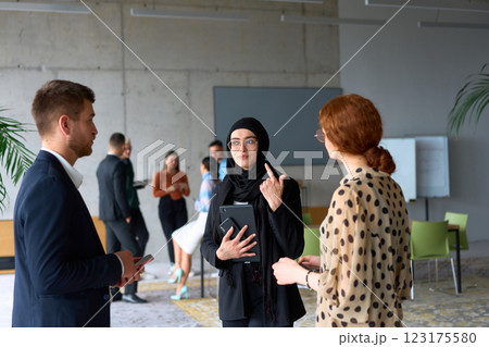 A diverse group of business professionals engages in a conversation during a work break in the office, with their colleagues visible in the background, fostering collaboration and connection 123175580