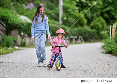A joyful teenager encourages her younger sister as she learns to ride a bike, both sharing a moment of laughter and bonding in the sunny outdoors 123175581