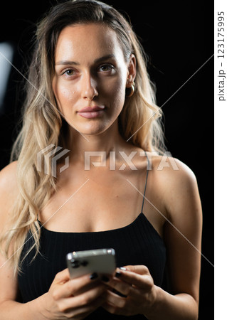 A blonde woman in a black dress posing elegantly in a studio with a black background, holding a smartphone in her hands A blonde woman in a black dress posing elegantly in a studio with a black background, holding a smartphone in her hands 123175995