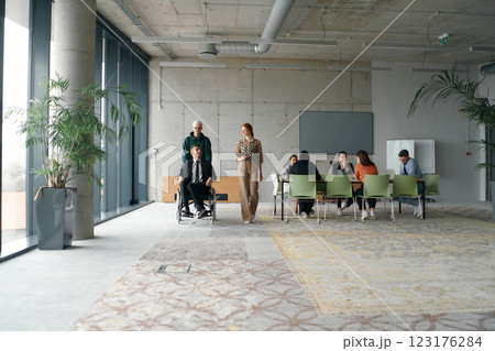 A diverse and multicultural business team collaborates in the office while their colleagues, including a director in a wheelchair and his friend, share a lighthearted moment, fostering an atmosphere 123176284