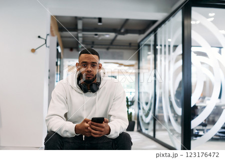 African American businessman wearing headphones while using a smartphone, fully engaged in his work at a modern office, showcasing focus, productivity, and contemporary professionalism African American businessman wearing headphones while using a smartphone, fully engaged in his work at a modern office, showcasing focus, productivity, and contemporary professionalism 123176472