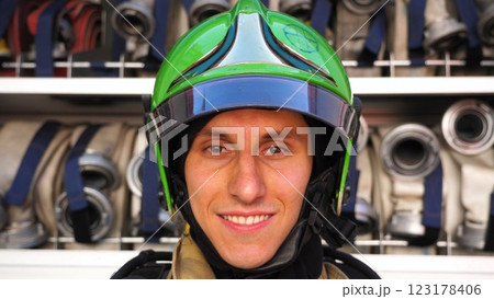 Young smiling fireman looking at camera in full equipment after fire against the background of big red truck. Portrait of happy male fireguard standing near a fire engine. Concept of heroic profession Young smiling fireman looking at camera in full equipment after fire against the background of big red truck. Portrait of happy male fireguard standing near a fire engine. Concept of heroic profession 123178406