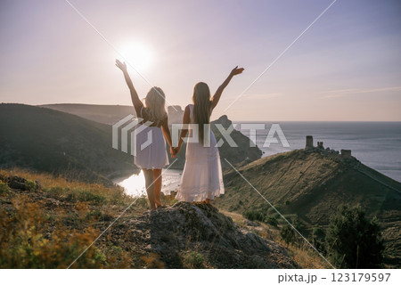 Two women are standing on a hill overlooking the ocean. They are holding hands and looking out at the water. The scene is peaceful and serene, with the sun shining brightly in the background. Two women are standing on a hill overlooking the ocean. They are holding hands and looking out at the water. The scene is peaceful and serene, with the sun shining brightly in the background. 123179597