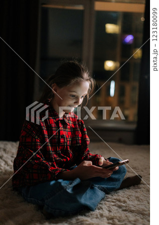 Vertical shot of pretty preteen browsing smartphone sitting on bed by window at night in dark bedroom, smiling scrolling social media with bokeh city lights glowing softly on blurred background. Vertical shot of pretty preteen browsing smartphone sitting on bed by window at night in dark bedroom, smiling scrolling social media with bokeh city lights glowing softly on blurred background. 123180099