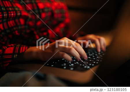 Close-up cropped shot of unrecognizable preteen girl wearing red and black checkered shirt typing on backlit laptop keyboard sitting on bed in dark room, creating cozy atmosphere. 123180105