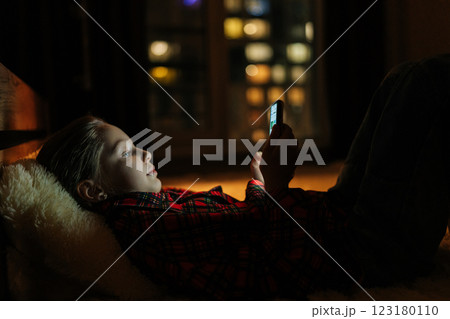 Closeup side view of relaxed young girl lying in bed at night, using smartphone illuminated by soft glow of screen, with blurred city bokeh lights twinkling outside window, creating cozy atmosphere. 123180110