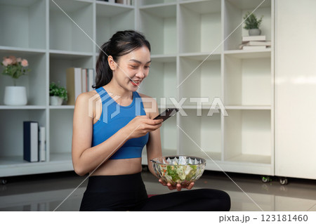 Woman sitting cross legged with a bowl of healthy salad and smartphone in a modern living space. 123181460