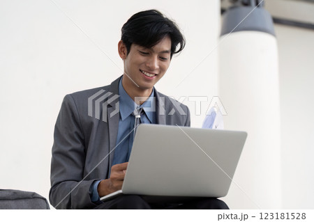Young Asian businessman smiling while working on a laptop in a modern outdoor business environment. Young Asian businessman smiling while working on a laptop in a modern outdoor business environment. 123181528