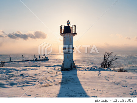 A white lighthouse stands tall on a snowy shore, casting a long shadow as the sun sets over the frozen sea 123185308