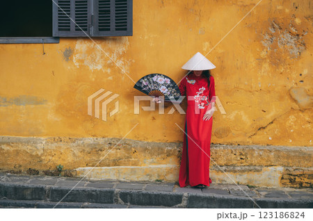 Asian young woman traveler tourist in traditional vietnamese non la hat and red dress with a fan on an old town street in Hoi An in Vietnam in Asia 123186824
