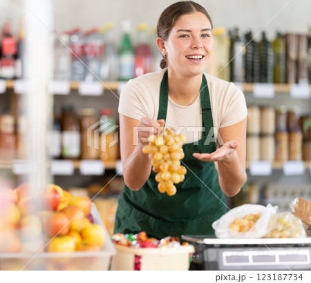 Young woman seller in store is standing near fruit counter, selling grape. Young woman seller in store is standing near fruit counter, selling grape. 123187734