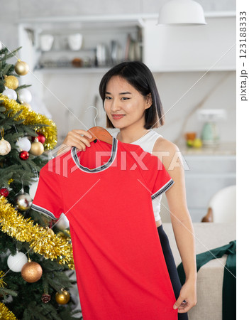 Happy young Asian girl trying to choose a dress standing next to Xmas-tree 123188333