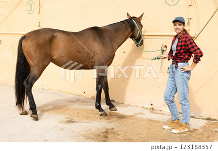 Young cowgirl farm worker washing horse off with hose on hot day in stable yard 123188357