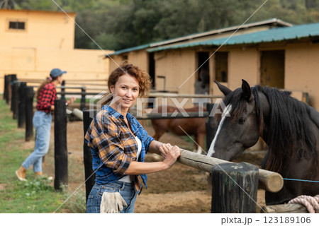 Portrait of positive smiling woman wearing plaid shirt and posing with horse on yard of horse stables Portrait of positive smiling woman wearing plaid shirt and posing with horse on yard of horse stables 123189106
