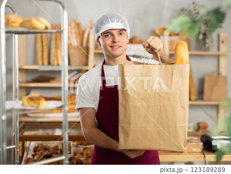 Portrait of positive young baker with paper bag of fresh baked goods in the interior of private bakery Portrait of positive young baker with paper bag of fresh baked goods in the interior of private bakery 123189289
