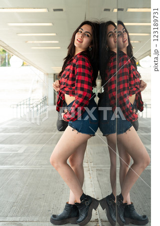 Stylish young woman posing against a glass wall, creating a mirrored reflection Stylish young woman posing against a glass wall, creating a mirrored reflection 123189731