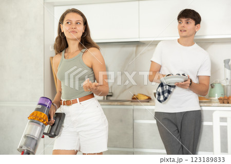 Young woman vacuum-cleaning in the kitchen while communicating with her boyfriend drying dishes with towel Young woman vacuum-cleaning in the kitchen while communicating with her boyfriend drying dishes with towel 123189833