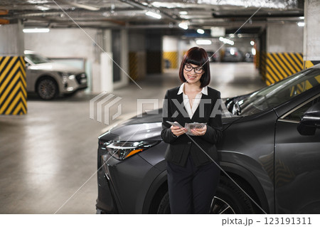 Caucasian businesswoman standing in parking garage, smiling while holding cash. Indoor setting with cars, showcasing finance, success, and confidence themes. Professional attire. 123191311