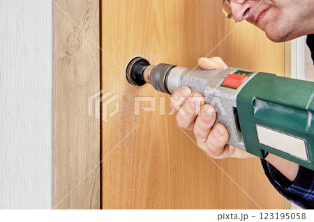 Man uses an electric drill with hole saw bit to create holes in door leaf for installing new door handle and latch. Man uses an electric drill with hole saw bit to create holes in door leaf for installing new door handle and latch. 123195058