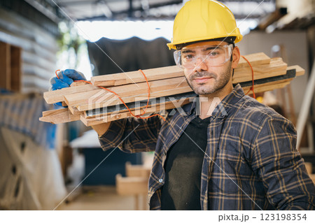 Smiling construction worker in a yellow hardhat and plaid shirt holding wood planks on his shoulder. Ideal for carpentry, woodworking, and craftsmanship in furniture workshop, National Carpenters Day 123198354