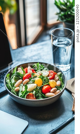 A healthy office lunch with bean salad, white beans, arugula, and cherry tomatoes, served on a desk beside a laptop and water glass. 123200040