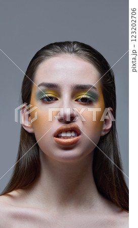 Close-up portrait of expressive young woman showing fierce expression with vibrant green and yellow makeup against gray background. 123202706