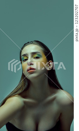 Young woman tilting head slightly while wearing bright green and yellow artistic makeup against teal background. 123202707