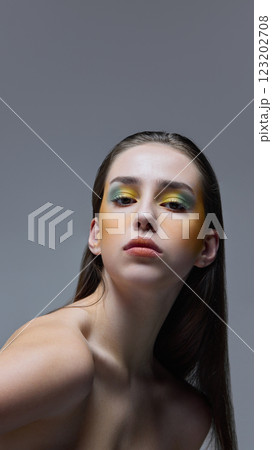 Artistic portrait of young, beautiful woman with green and yellow artistic makeup gazing confidently against soft gray background. Artistic portrait of young, beautiful woman with green and yellow artistic makeup gazing confidently against soft gray background. 123202708