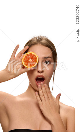 Young woman holding fresh orange slice over eye, and looking at camera with surprised expression against white background. 123202746