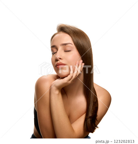 Portrait of young brunette woman with long smooth hair resting chin on hand with relaxed expression against white studio background. 123202813