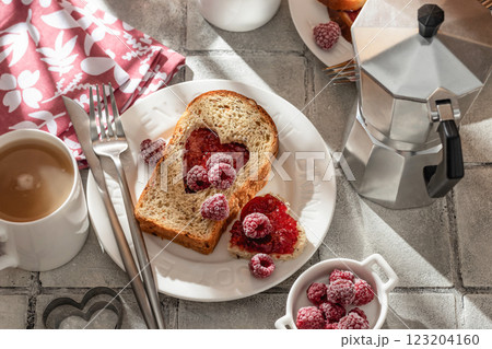 Toasted bread with heart-shaped cut and raspberry jam on tile background in bright morning light, top view. Festive breakfast for Valentines day 123204160