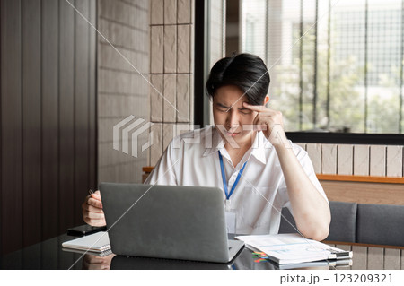 Young Asian male office worker experiencing stress while working on laptop, focusing on problem-solving and analysis. Young Asian male office worker experiencing stress while working on laptop, focusing on problem-solving and analysis. 123209321