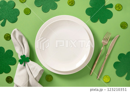 St Patrick's Day table setting with white plate and traditional decoration, shamrock, clover leaves and coins on light green background. View from above. 123210351
